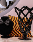 Black decorative vase with intricate lattice design on a white surface with a black bow and orange and black striped pillow in the background.