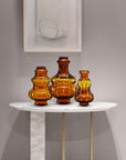 Three amber-colored crystal vases on a white table against a gray wall with a framed picture.