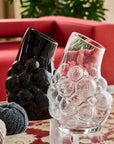 Two crystal vases, one black and one clear with bubble design, on a table with a red couch and plant in the background.