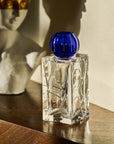 Clear crystal vase with blue top on a wooden surface, with a blurred statue in the background.