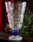 Decorative crystal vase with a blue base on a dark surface with a blurred green plant and red curtain in the background.