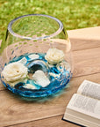 Crystal vase with blue water and white flowers on a wooden surface with an open book.