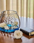 Crystal vase with white flowers and blue water styled on a table next to a small book. 