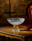 Clear crystal bowl with gold base on a wooden table against a stone wall.