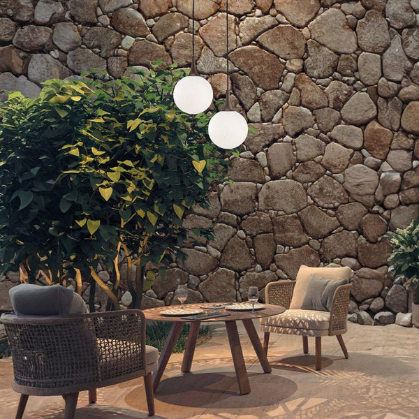 Outdoor seating area with round pendant lights from Masiero, wicker chairs and a wooden table against a stone wall.