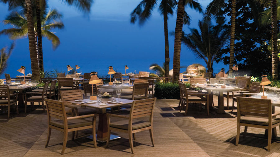 Outdoor dining area with tables and chairs under palm trees at dusk and Masiero's Cordea rechargeable table lamps on each table. 