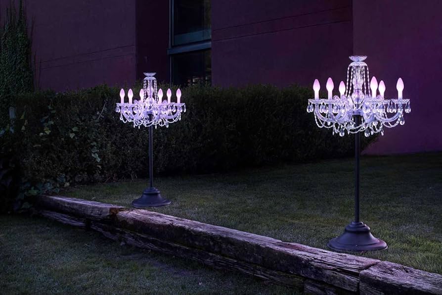 Two illuminated chandelier lights on stands in a garden setting with a dark background.
