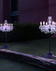 Two illuminated chandelier lights on stands in a garden setting with a dark background.