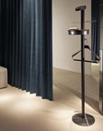 Black coat stand with a round base and a rectangular top in a room with dark curtains and tiled wall.