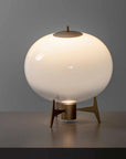 A modern table lamp with a large, glowing frosted glass globe shade on a sculptural bronze-colored metal tripod base, set against a dark gray background.