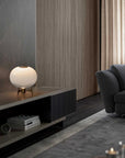 A modern spherical white table lamp with a warm brass base sits on a dark floating TV console in a contemporary living room setting.