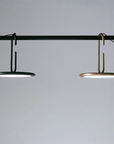 Two modern circular pendant lights hanging from a black horizontal rod against a light gray background. One light has a black frame and the other has a warm gold/brass frame, both emitting a soft glow.
