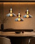Three modern pendant lights with dark bronze metal shades and glowing warm yellow bulbs, hanging over a round wooden dining table in a warmly lit contemporary dining room.