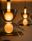 A close-up of a modern pendant light with a translucent amber-tinted glass shade and a glowing warm white bulb, shown against a softly blurred interior background with additional matching lights in the distance.