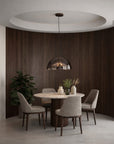 A modern dining room setup featuring a round light-wood table surrounded by five upholstered dining chairs, with a dome-shaped pendant light hanging above. The room has a curved dark wood-paneled wall, a light gray concrete floor, and a recessed ceiling light.