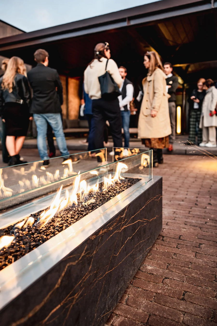 People gathered around a fire pit on a brick patio with a building in the background