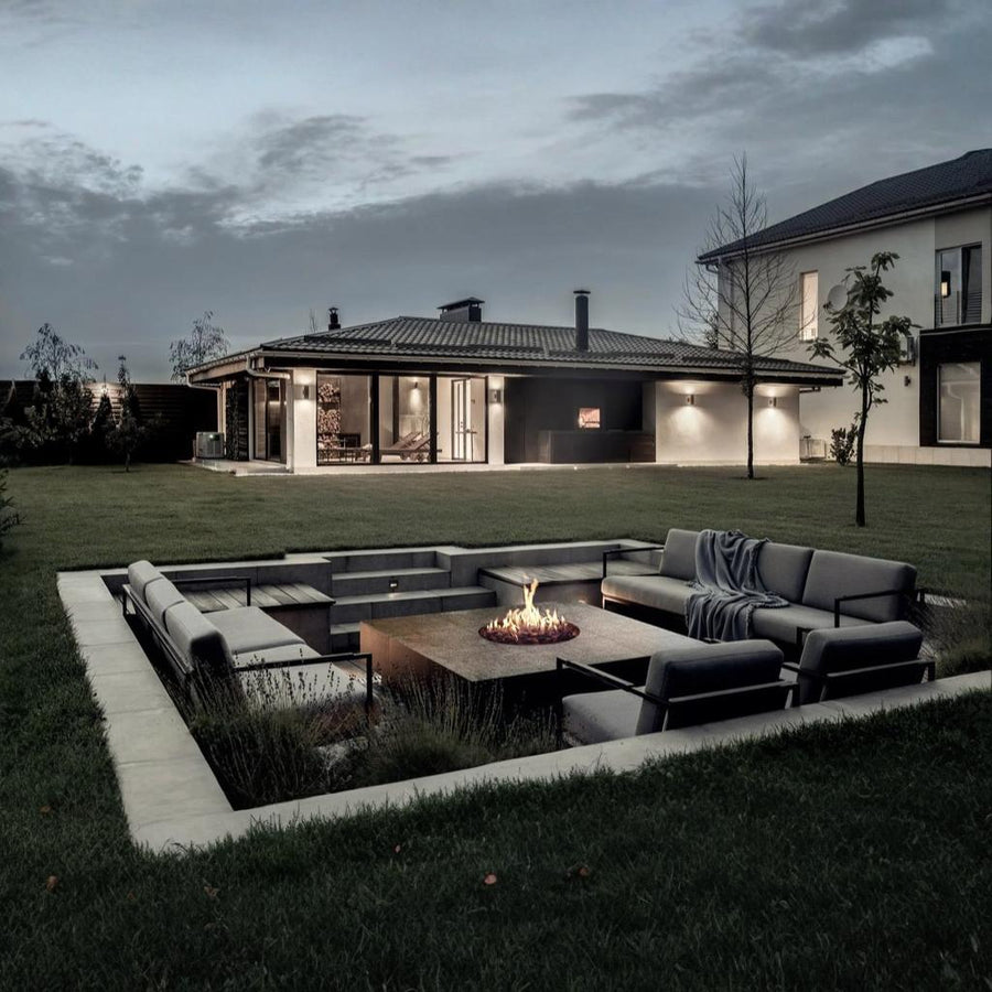 Modern outdoor seating area with a gas fireplace in a garden setting at dusk.