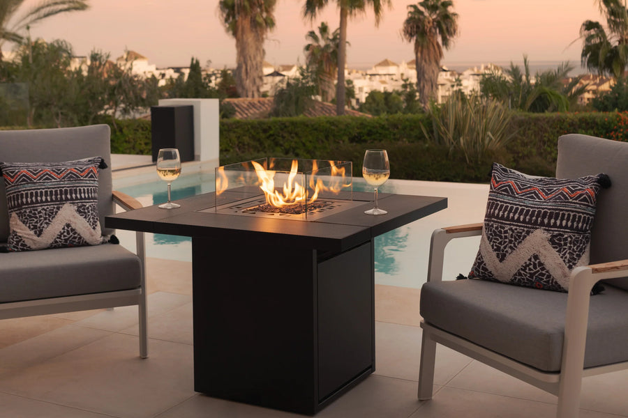 Outdoor patio setup with fire pit table, chairs, and decorative pillows by a pool.