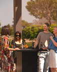 Four people standing around a gas fireplace table with drinks outdoors, surrounded by greenery.