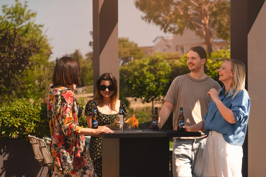 Four people standing around a gas fireplace table with drinks outdoors, surrounded by greenery.