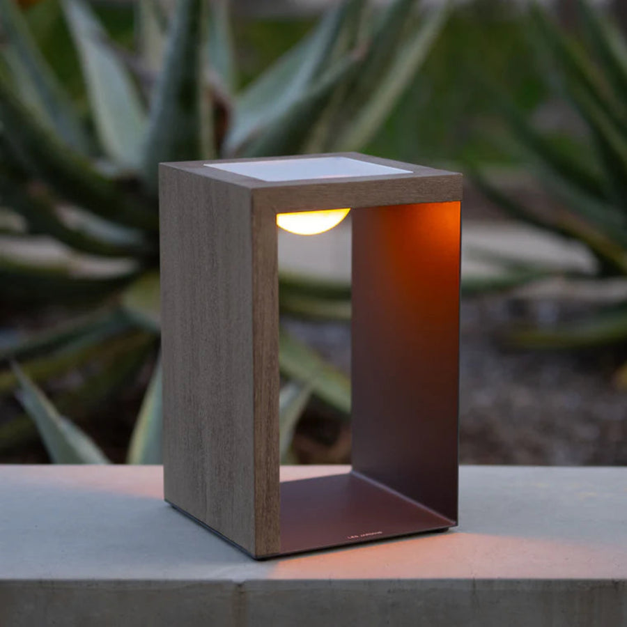 Modern outdoor solar lantern with wooden finish on a concrete surface, blurred green plants in the background by Les Jardins available at Spacio retail stores in India.