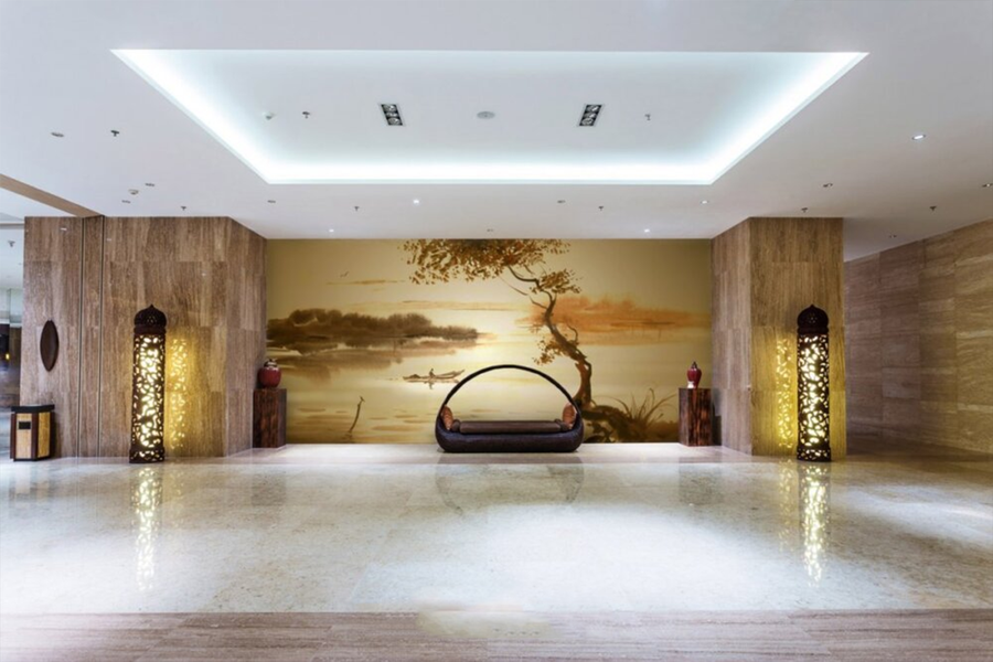 Golden watercolour fresco mural depicting a calm lake, a small boat, and a windswept tree, handcrafted on Italian plaster with Carrara marble and travertine powder. Featured on a wall in a hotel lobby in front of a modern design basket sofa 