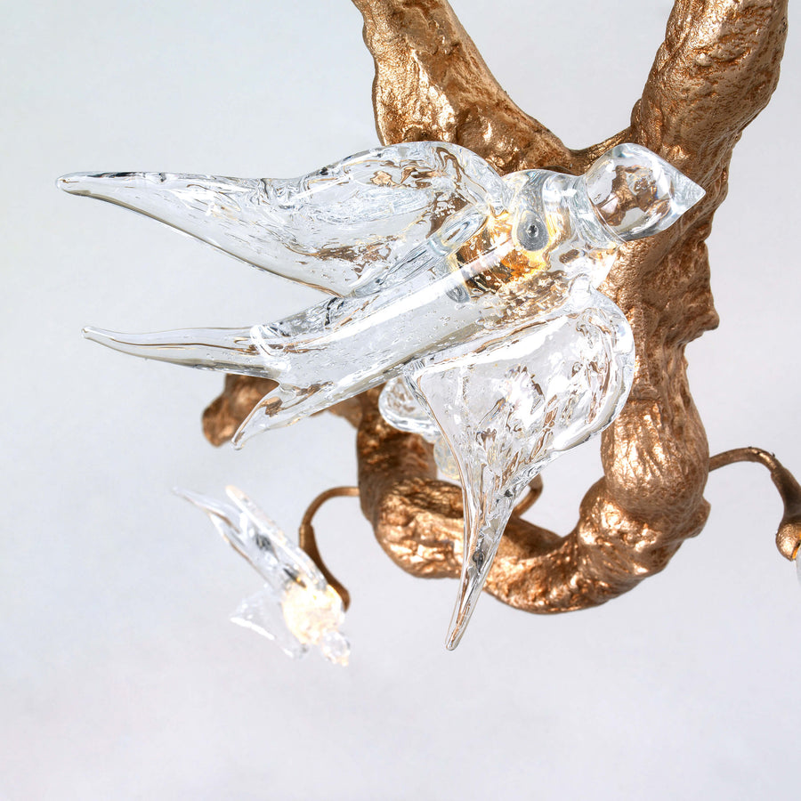 Close up of a chandelier with glass bird sculptures on a gold antler against a light gray background