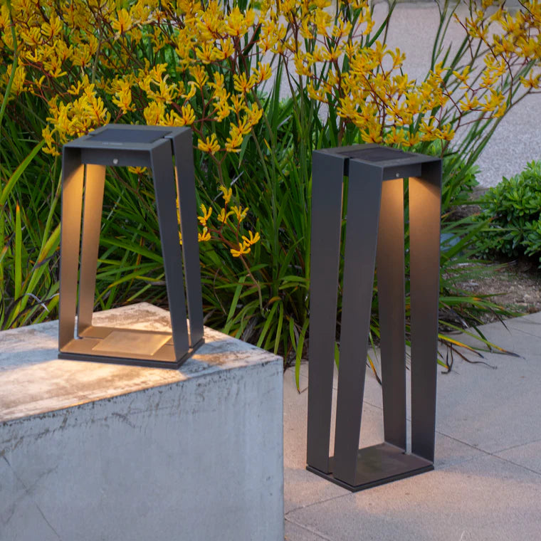 Two modern outdoor lamps on a stone ledge with yellow flowers in the background by Les Jardins available at Spacio retail stores in Mumbai & Kolkata. 