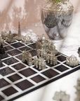 A close-up of miniature Indian monuments   architectural Skyline chess game in progress, featuring silver and black city-piece towers on a dark grid board, set on a marble surface with a blurred glass vase in the background.