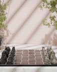 A decorative Indian monuments Skyline chess set displayed on a marble surface, featuring black and silver castle-shaped chess pieces on a dark board. The scene is styled with potted green plants and soft diagonal shadows on a blush pink background.