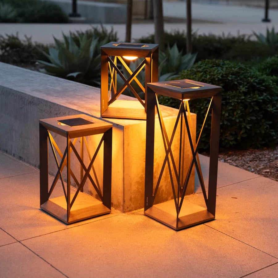 Three illuminated outdoor lanterns on a stone surface with plants in the background by Les Jardins available at Spacio retail stores in Mumbai & Kolkata. 
