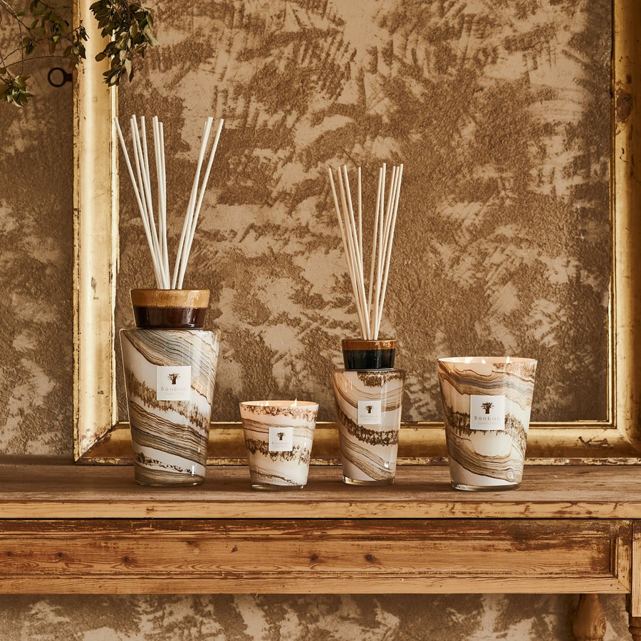 Four hand-blown glasses holding Baobab Sand Siloli Candle MAX16SSI fragrance diffusers are placed on a wooden table, complemented by a Baobab candle.