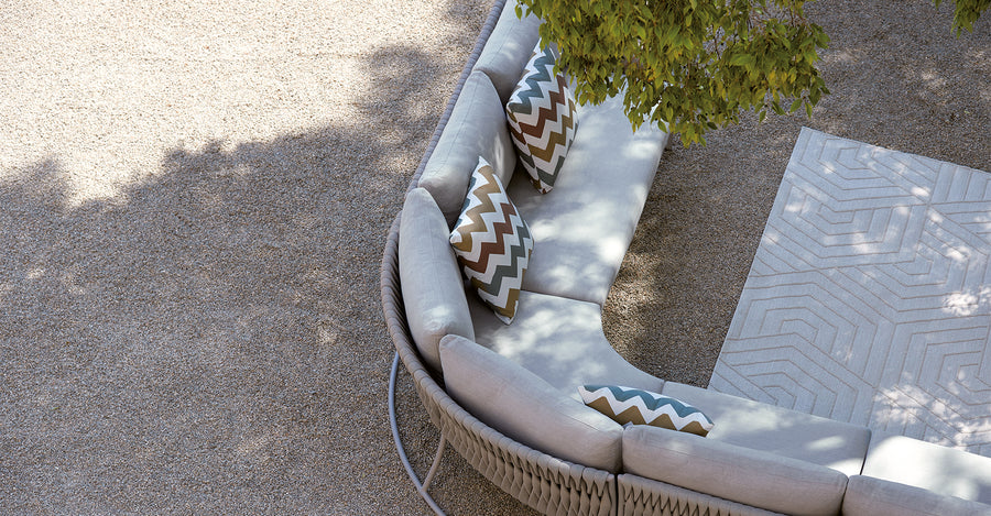 The Sifas Basket Sofa Collection, designed by Sifas, is an outdoor furniture basket collection featuring a white couch with pillows. It is specifically designed with waterproof properties for outdoor use. The couch is placed against a backdrop of a majestic tree in the background.