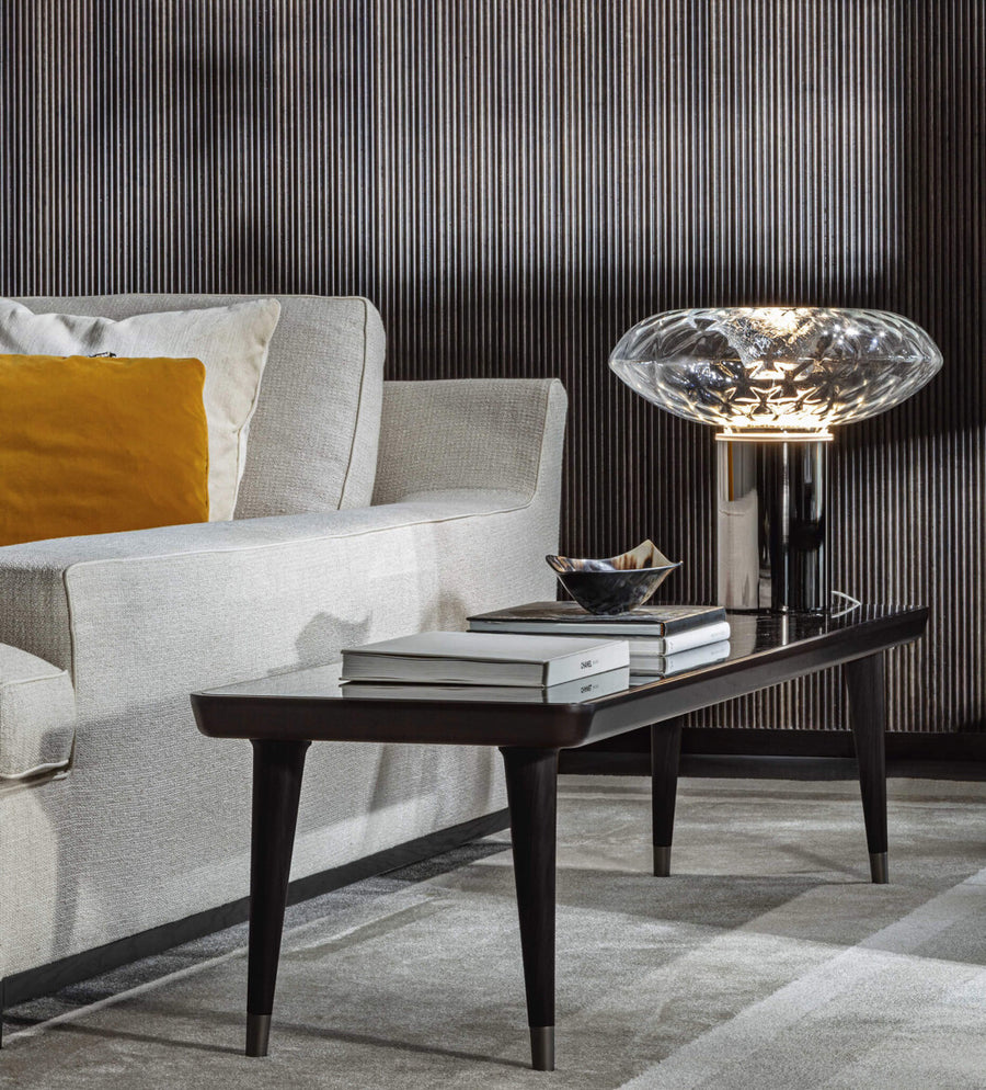 A table lamp on a marble side table next to a sofa with an orange pop colour cushion in a luxury living room with a dark theme. Cicla collection by Italamp, featuring handmade Balloton glass lamps in transparent, white, and smoked finishes with grooved Vicenza stone bases and metal accents.