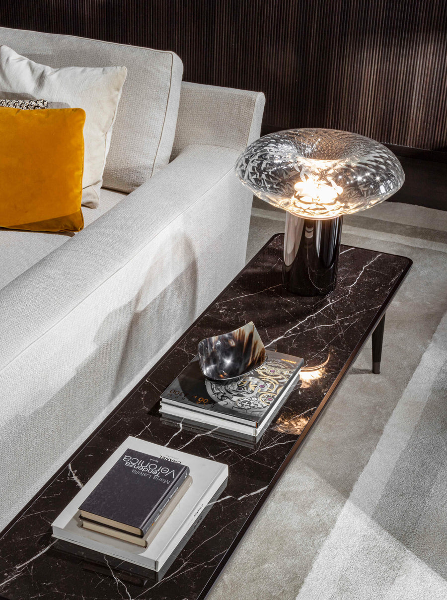 A table lamp on a marble long side table next to a sofa in a contemporary theme living room styled along with coffee table books and art objects. Cicla collection by Italamp, featuring handmade Balloton glass lamps in transparent, white, and smoked finishes with grooved Vicenza stone bases and metal accents.