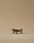 A small sculptural tabletop bowl with a shallow oval bowl, three short legs, and dark side details, photographed against a seamless warm beige studio background.