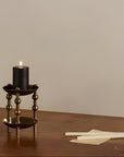 A black pillar candle lit in a brass two-tiered candle holder, set on a wooden table with unlit white candles nearby against a neutral beige background.