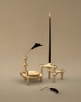 A minimalist set of brass candlestick holders and vessels with a tall dark taper candle, a small black flower, and a loose flower stem, arranged on a warm beige studio background.