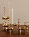 A set of modern brass candlesticks and small holders arranged on a dark wooden table against a warm beige wall, with two lit white taper candles placed among them.