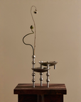 A modern metallic three-tier candle holder with spherical bead details on its legs, styled with a single dried flower stem, displayed on a dark wooden pedestal against a warm beige background.