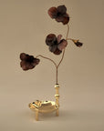 A decorative gold-toned metal candle holder with a vertical stem and three dark brown flower-like ornaments, shown against a plain warm beige background.