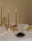 A set of ornate gold brass candlesticks with three taper candles lit, arranged on a white tablecloth alongside a plate of pears, a bowl of dark cherries, and a cream ribbon against a warm beige background.