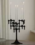 A black metal multi-arm candelabra holding five white taper candles, lit and set on a wooden surface against a cream-colored wall with a window sill and curtain in the background.