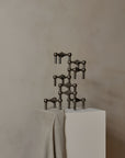 A dark metallic candle stand made of linked angular forms and small spherical elements, displayed on a white pedestal draped with a light neutral cloth against a warm beige wall.