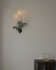 A black metal wall sconce with five candle-style arms, shown lit against a plain beige wall above a dark wooden shelf with a glass vase and books.