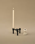 A black metal candlestick holder with a single cream candle inserted, shown next to a small white decorative object on a warm beige studio background.