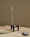 A black metal candle holder with a single lit white LED candle on a wooden table, with a small remote control lying nearby against a warm beige background.
