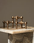 A bronze geometric candle stand sculpture with spherical and conical forms displayed on a polished beige marble pedestal against a warm taupe background.