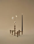 A sculptural dark brown metal candle holder with geometric supports, holding a black taper candle and three small white flowers, set against a solid warm beige background.
