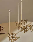 A set of modern brass candlesticks with bulbous geometric arms holding three tall white taper candles, arranged on a beige table next to a brown ceramic mug against a warm beige background.